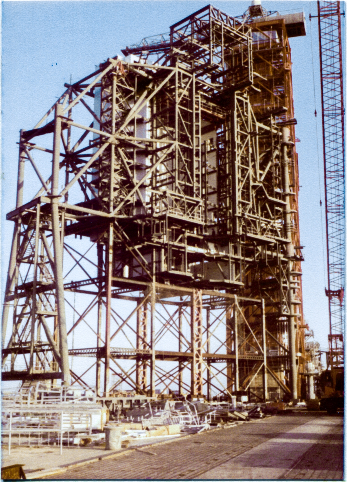 Image 007. Rising nearly 200 feet above the level of the Crawlerway you’re looking at it from (which itself rises nearly 50 feet above the level of the surrounding wetlands), the Rotating Service Structure at Space Shuttle Launch Complex 39-B, Kennedy Space Center, Florida, stands as an open skeleton, still partially supported on the falsework which the Union Ironworkers working for Wilhoit Steel Erectors constructed first, to hold it up temporarily while it was being erected, before it became self-supporting and the falsework could then be removed. Behind the gray steel of the RSS, the red framework of the taller Fixed Service Structure, which was built first, reusing parts of an Apollo Project Launch Umbilical Tower, can also be seen. On the pad deck in the foreground beneath the falsework, additional material fabricated by Sheffield Steel, who fabricated all of the structural steel for the RSS, can be seen, awaiting its turn to be lifted into place by Wilhoit’s big Manitowoc crane, visible along the right margin of the image. Photo by James MacLaren.